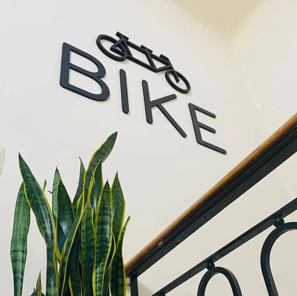 Logo de BIKE en la pared de la escuela Logo en relieve de la escuela de inglés BIKE sobre una pared blanca con plantas decorativas y baranda en primer plano.